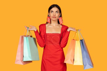 Photo for A woman dressed in a chic red dress stands against a warm yellow background, her hands holding several shopping bags in various pastel shades. Bright red sale tags are attached to her ears - Royalty Free Image