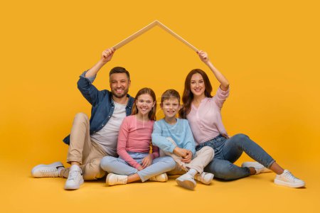 Eine glückliche Familie bestehend aus Vater, Mutter, Tochter und Sohn sitzt zusammen auf gelbem Grund und lächelt, während sie einen weißen Bilderrahmen über ihren Köpfen halten.
