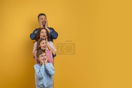 Téléchargez les photos : Famille surprise de quatre, avec deux enfants et leurs parents, est aligné verticalement, chacun plaçant leurs mains sur leurs joues dans une expression ludique de surprise et de bonheur, espace de copie - en image libre de droit