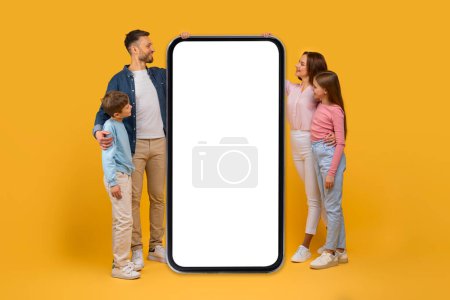 Téléchargez les photos : Une famille composée d'un homme, une femme, un garçon et une fille se tiennent ensemble à côté d'un smartphone surdimensionné avec un écran blanc vierge, adapté à des fins de maquette. - en image libre de droit