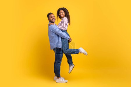 A joyful African American man is holding a woman in his arms as they both smile radiantly. They are dressed in casual clothing and seem to be engaged in a playful moment.