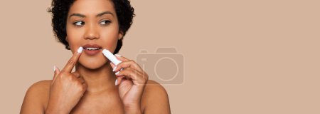A young woman with curly hair and a thoughtful expression applies lip balm carefully to her lips, standing against a cohesive beige backdrop that accentuates her complexion, copy space