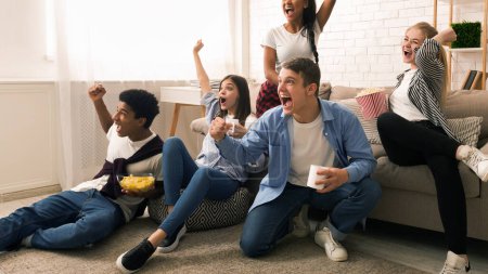Un grupo de amigos se reúne en una acogedora sala de estar, expresando alegría y emoción. Algunos están sentados en un sofá mientras que otros se sientan en el suelo, viendo un partido de deportes o un evento competitivo en la televisión..