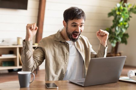 Photo for A young man seated at a wooden desk raises his fists in elation, his face expressing excitement and joy, as he looks at his laptop screen. A smartphone and a coffee cup rest nearby - Royalty Free Image