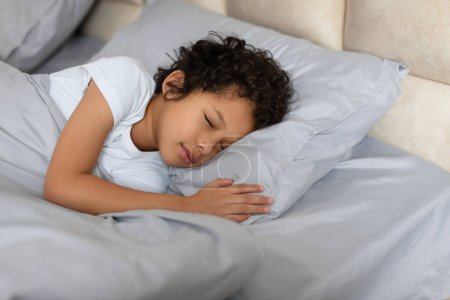 A small African American child peacefully sleeping on a bed while hugging a fluffy pillow. The child is nestled in blankets, and their eyes are closed in deep slumber.