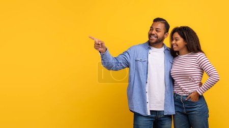 Ein afroamerikanisches Paar zeigt auf ein unsichtbares Objekt, während es vor einem leuchtend gelben Hintergrund steht. Beide wirken engagiert und fokussiert auf das, was sie andeuten, den Raum kopieren