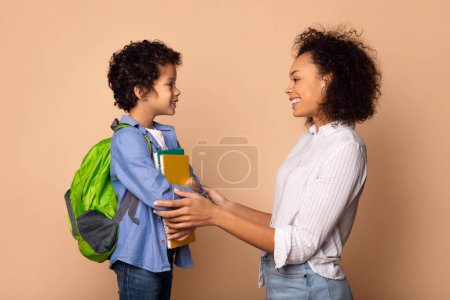 Un joven afroamericano, equipado con una mochila verde y sosteniendo libros coloridos, se para frente a su madre sonriente mientras le ofrece apoyo y afirmación antes de que se vaya a la escuela.