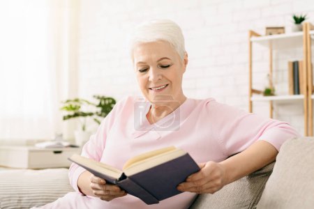 Senior woman is seated on a couch, engrossed in reading a book. She is focused on the pages, holding the book in her hands.