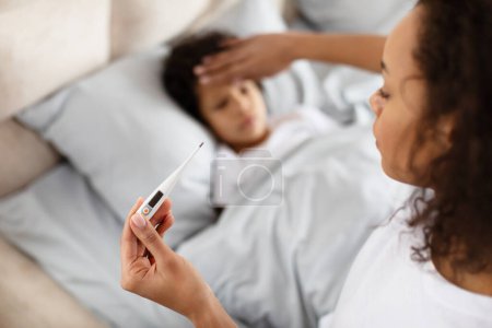 A caring African American mother is gently placing her hand on her child forehead to feel for a fever while holding a digital thermometer. The child appears to be resting in bed