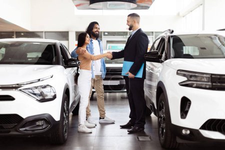 A professional car salesman, dressed in a suit, is shaking hands with a happy Indian couple customers inside a car dealership showroom