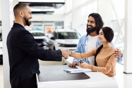 Photo for A man and a woman are shaking hands inside a well-lit car showroom. The man appears to be a car salesman, and Indian woman is a potential customer. They are standing next to a shiny car - Royalty Free Image