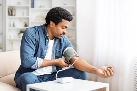 A focused young African American man sits comfortably on a couch while he wraps a blood pressure cuff around his upper arm, monitoring his blood pressure in the relaxed atmosphere of his living room.