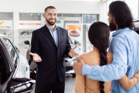 A cheerful car salesman is having a conversation with Indian couple inside a car dealership showroom, he seems to be explaining the features or benefits of a vehicle
