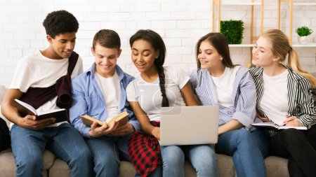 Five multiethnic teenagers are sitting closely together, sharing a casual study session indoors. Two of them are reading books, one is typing on a laptop, and the others appear to be discussing