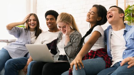 A diverse group of five friends is seated closely on a couch, sharing a fun moment as they look at a laptop screen together.