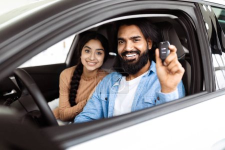 A cheerful and excited Indian couple is seated inside a new car. The man, with a beard, holds up car keys, indicating they have likely just purchased the vehicle, while the woman smiles warmly