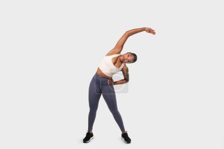 Photo for A young African American woman is captured in a moment of fitness, performing a side stretch exercise. She is wearing athletic attire, consisting of a white tank top, grey leggings - Royalty Free Image