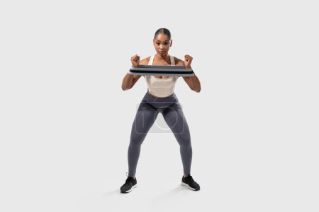 A focused African American woman is engaging in a fitness routine using a resistance band to strengthen her upper body muscles. She maintains a squat position, highlighting her athletic form