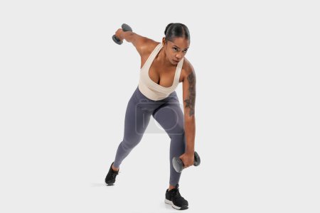 Téléchargez les photos : Femme afro-américaine s'exerce avec des haltères sur un fond blanc uni. Elle est concentrée et déterminée, soulevant les poids avec précision et contrôle. - en image libre de droit