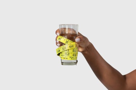 Foto de Recortado de mujer negra se muestra sosteniendo una cinta métrica hasta un vaso de agua, posiblemente midiendo la altura o el volumen del líquido, fondo blanco - Imagen libre de derechos