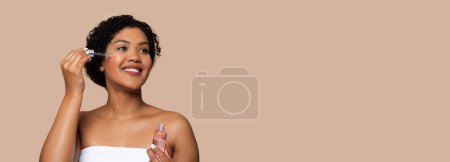 Photo for A smiling young woman with her hair up is applying a clear facial serum to her cheek using a dropper. She is wrapped in a white towel, indicating morning routine, panorama, copy space - Royalty Free Image