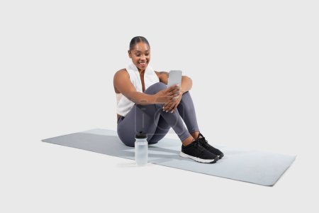 Black woman with a bright smile rests on a yoga mat, dressed in workout clothes, and enjoys a break from her exercise routine in a brightly lit indoor space, using phone