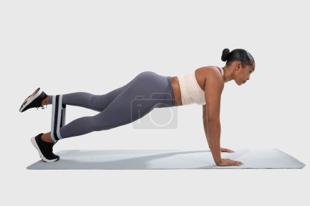 Téléchargez les photos : Femme afro-américaine effectue push-ups sur un tapis dans une salle de gym ou un studio de fitness. Elle engage ses muscles du haut du corps tout en maintenant une position de planche, en utilisant une bande de résistance - en image libre de droit