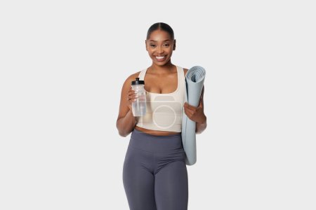 Téléchargez les photos : Femme afro-américaine debout tout en tenant un tapis de yoga dans une main et une bouteille d'eau dans l'autre. Elle semble prête pour une séance d'entraînement ou un cours de yoga, avec une expression ciblée. - en image libre de droit