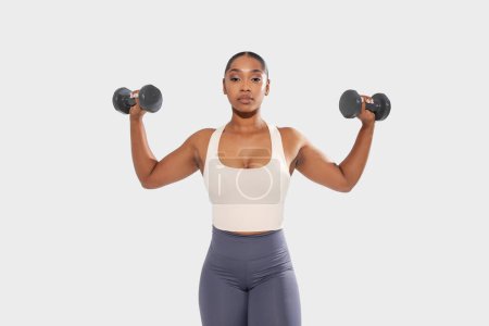 African American woman is shown in the image holding two dumbbells in her hands. She is in a gym setting, demonstrating strength and fitness by engaging in weightlifting exercises.