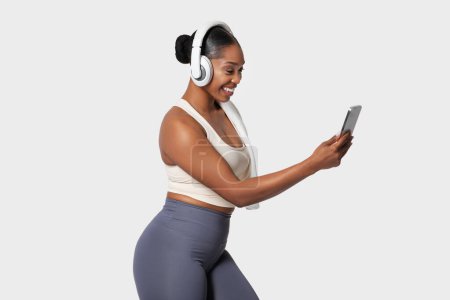Foto de Una alegre mujer afroamericana está comprometida con el teléfono, con un par de auriculares para la oreja, vestida con un atuendo casual de entrenamiento. Su atención se centra en la pantalla - Imagen libre de derechos