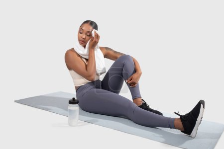 Eine junge Afroamerikanerin in Trainingsausrüstung sitzt mit einem Bein auf einer Yogamatte und stützt ihren Ellbogen auf das gebeugte Knie ihres anderen Beines mit einem Handtuch ab.
