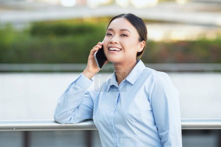 An Asian businesswoman is engaged in a phone conversation, conveying professionalism and focus. She is seen multitasking while maintaining a business demeanor