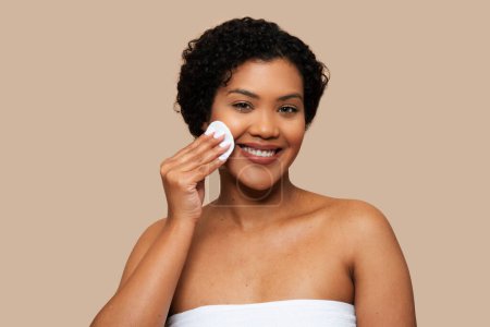 A cheerful young woman wrapped in a white towel is gently applying skincare product to her face using a cotton pad. Her natural curly hair, clear skin, and bright smile convey a sense of beauty