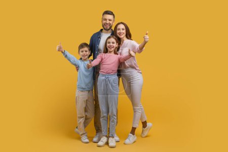 Happy family consisting of a mother, father, son, and daughter stands close together, all giving a thumbs up sign. They are posing against a vivid yellow backdrop