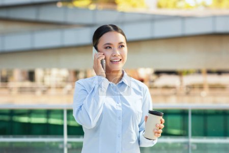 Foto de Una mujer asiática vestida con ropa de negocios se dedica a una conversación telefónica. Ella sostiene un teléfono inteligente en su oído mientras habla y escucha atentamente - Imagen libre de derechos