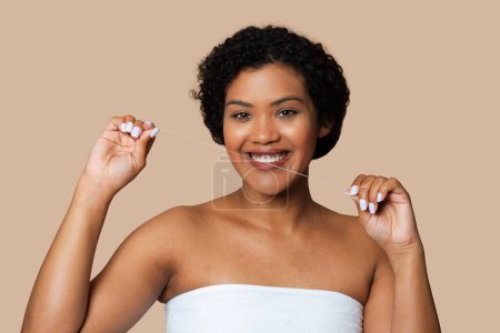 A young woman stands against a soft beige backdrop, smiling as she demonstrates proper dental floss usage with a length of floss between her fingers