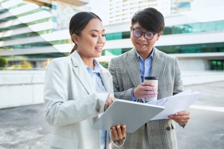 Two colleagues, Asian woman holding a tablet and a man with a coffee cup, are standing outside of a modern building complex, reviewing and discussing documents together with engaged expressions
