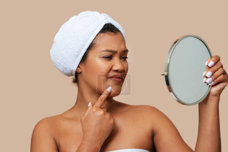 A woman is standing in front of a mirror, closely examining her own face. She appears focused and engaged, likely inspecting her skin or features. The mirror reflects her image back at her.