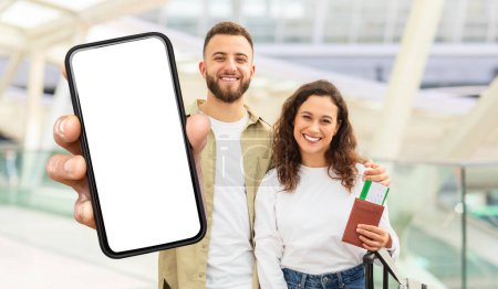 Una pareja radiante se encuentra en una terminal del aeropuerto, el hombre sostiene un teléfono inteligente con una pantalla en blanco hacia la cámara, mientras que la mujer a su lado agarra un pasaporte y una tarjeta de embarque.