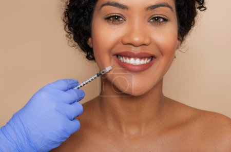 A young woman smiles gently as a healthcare professional wearing blue gloves administers a cosmetic injection near her mouth, suggesting a dermatological or aesthetic treatment.