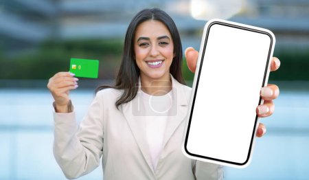 Foto de Una mujer está de pie y sostiene un teléfono inteligente en una mano y una tarjeta de crédito en la otra mano. Ella parece estar involucrada en una transacción o haciendo una compra.. - Imagen libre de derechos