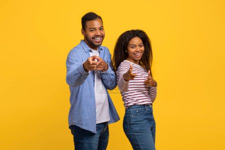 Un joven y alegre hombre y mujer afroamericanos se paran uno al lado del otro, sonriendo y señalando directamente al espectador con confianza y entusiasmo