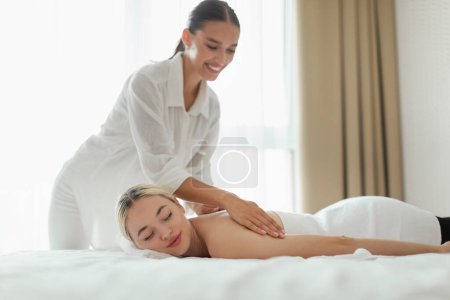 Foto de Una mujer está acostada boca abajo en una mesa de masaje mientras otra mujer, vestida con una camisa blanca, le está dando un masaje de espalda. El entorno parece ser un estudio de masaje profesional. - Imagen libre de derechos