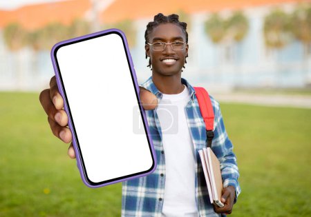 Un joven negro alegre con el pelo trenzado que lleva una mochila roja, sostiene un teléfono inteligente con una pantalla en blanco mientras está de pie en un campus universitario con edificios en el fondo