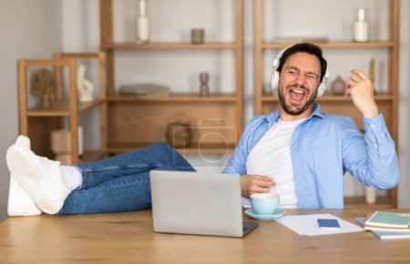 A delighted man is reclining with his feet up on a desk in a home office setting, wearing casual clothes and headphones, expressing joy while possibly taking a break from work