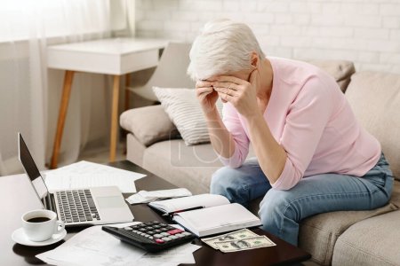 Eine ältere Frau sitzt auf einer Couch, die Hände verdecken ihr Gesicht und zeigen Anzeichen von Stress oder Kopfschmerzen, umgeben von Finanzunterlagen, einem Taschenrechner, Geld und einem Laptop.