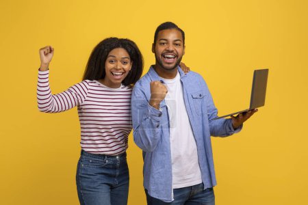 Photo for Black couple standing side by side against a yellow backdrop, both exuding excitement and joy. The man holds a laptop in one hand while they both raise their other fists in a triumphant gesture. - Royalty Free Image