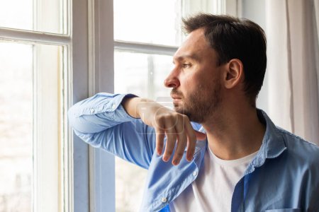 A man rests his chin on his hand as he thoughtfully looks out of a window, with gentle light filtering in, illuminating his casual attire and offering a peaceful ambiance to the start of his day.
