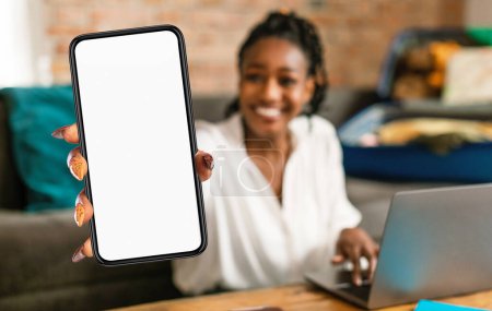 Photo for Black woman is seen holding up a cell phone in front of a laptop screen. She appears to be engaged in a video call or browsing content on the phone while using the laptop simultaneously. - Royalty Free Image