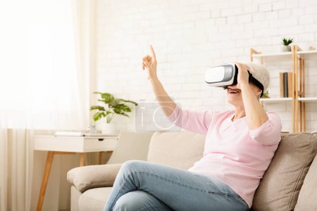 Une femme âgée est assise sur un canapé, portant un casque de réalité virtuelle, engagée dans le monde virtuel, avec une main tendue devant elle. La pièce est faiblement éclairée, avec le casque bleu brillant.